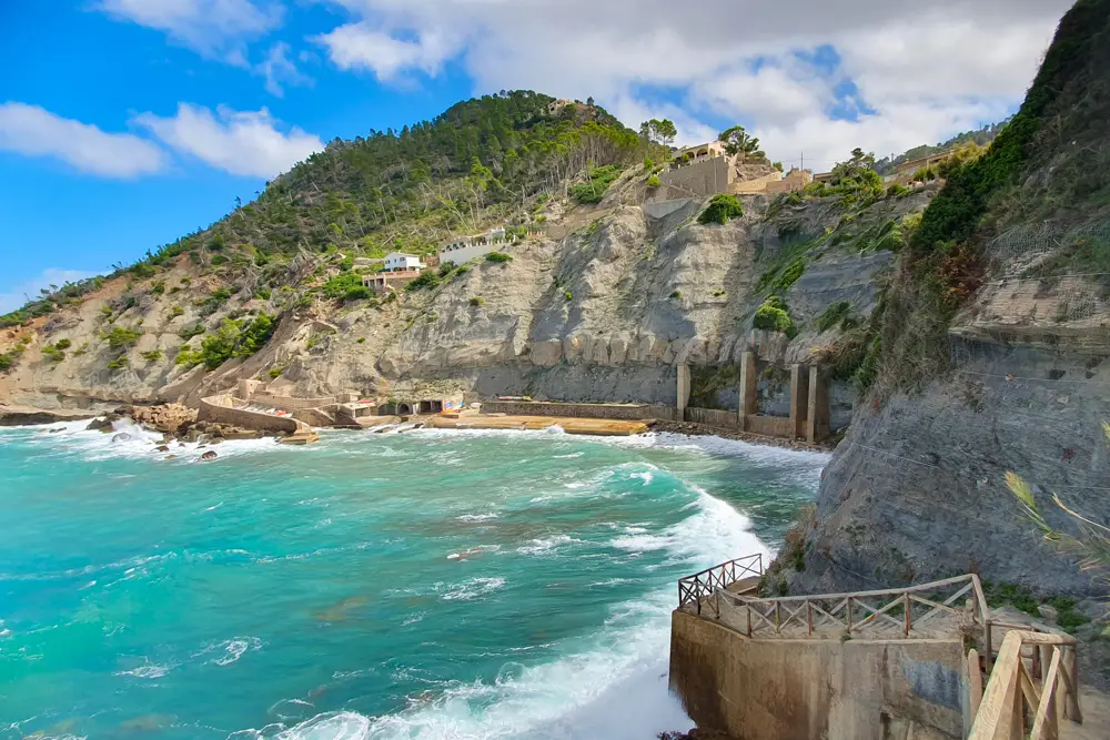 Ein malerischer Küstenblick auf steile Klippen in Banyalbufar, Mallorca, mit türkisfarbenen Wellen, die gegen das felsige Ufer schlagen. Der Hang ist mit terrassierter Vegetation, Steingebäuden und einigen Häusern nahe der Kante bedeckt. Ein hölzernes Geländer führt hinunter zum Wasser und verstärkt den rauen, idyllischen Charme der Mittelmeerküste.